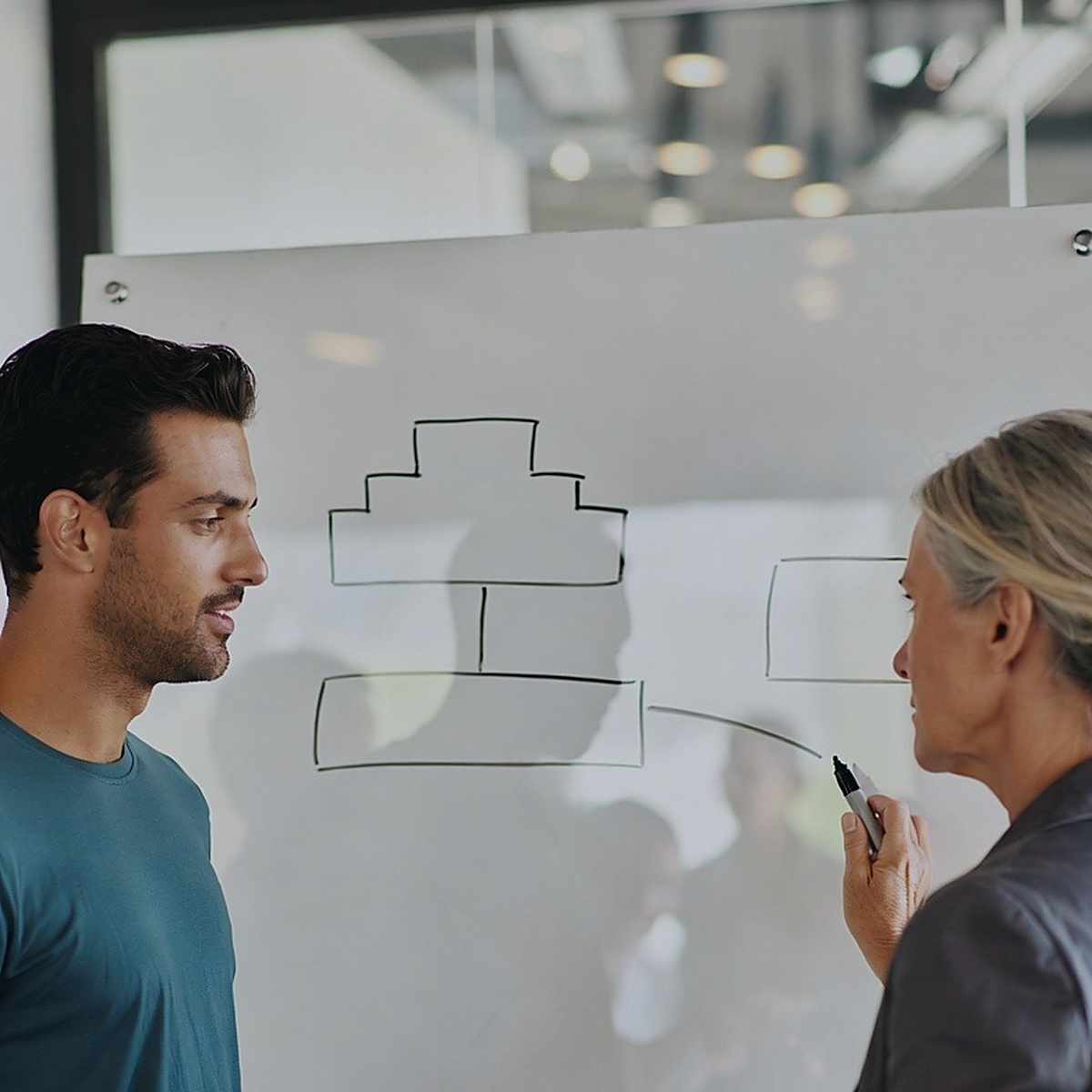Two colleagues at a whiteboard of tiered boxes and connectors, one holding a capped marker against the board, a glass wall onto an adjacent populated room behind them.