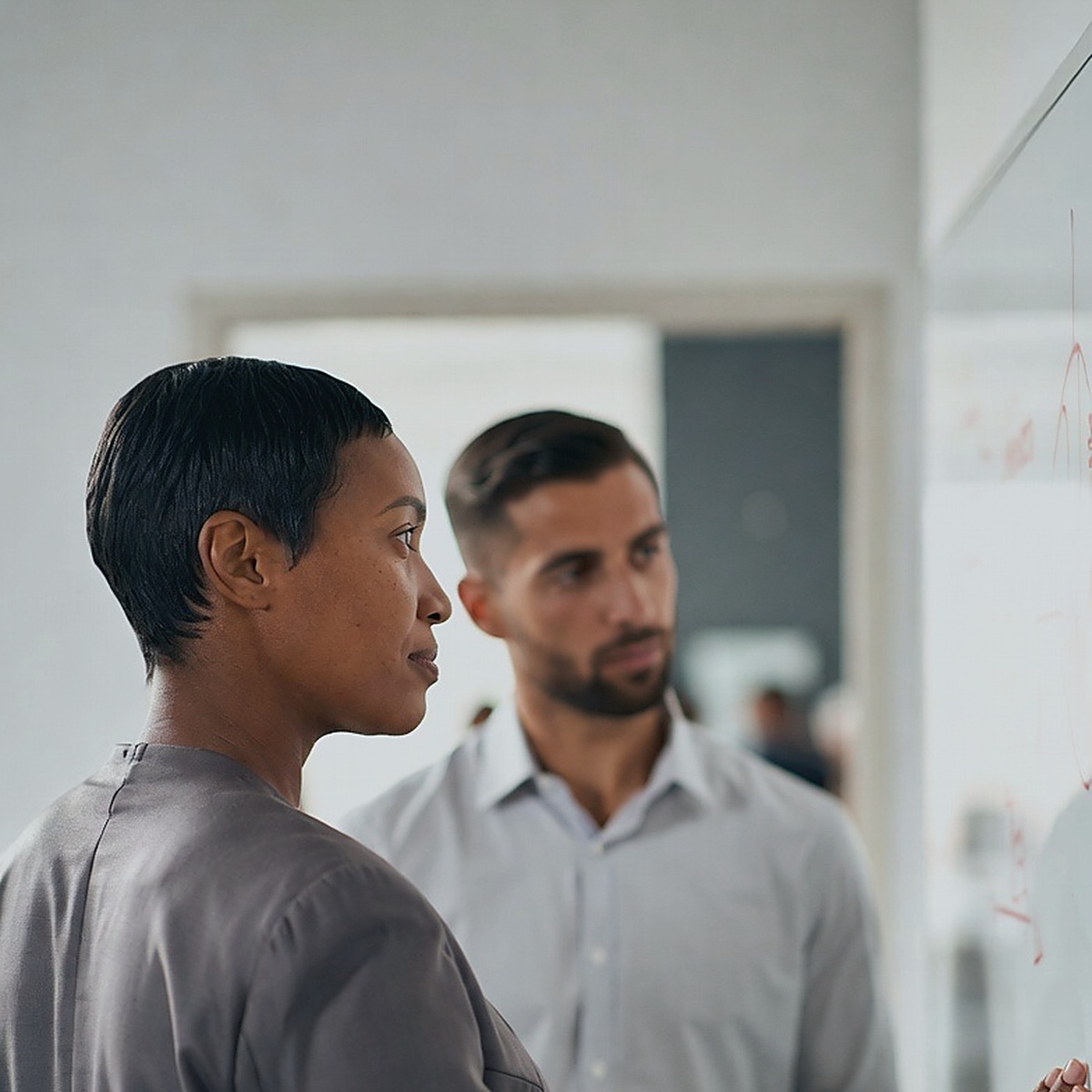 Two colleagues at a whiteboard, one indicating a point on the board while the other follows, a populated office floor visible through the doorway behind them.